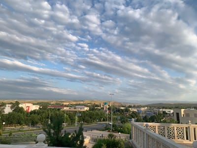 This is a high-angle, eye-level, wide shot of a city, likely Zaamin, Uzbekistan, under a partly cloudy sky. The foreground features a decorative stone railing and balustrade of a building, suggesting a vantage point from a terrace or elevated structure. Below, a road with a traffic lane and pedestrian walkway runs through the scene. Several cars are visible on the road, and a few individuals are walking on the sidewalk. Dense green trees line the roadside, with a large building featuring a red and white facade and a circular logo to the left. In the mid-ground, a mosque with blue domes and minarets stands prominently. Further in the distance, a range of rolling, arid hills forms the horizon, under a sky filled with elongated, striated clouds and patches of blue. The lighting suggests it is either early morning or late afternoon, with the sun casting a warm glow on the landscape.