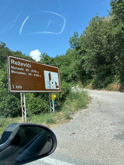 The image shows a brown road sign with white text and a white symbol. The sign reads "Reževići", "Manastir XII - XVIII v.", "Monastery XII - XVIII c.", and "1 km". The symbol on the sign depicts a stylized building that appears to be a church or monastery. The sign is mounted on a metal post and is surrounded by lush green trees and foliage. The sky is bright blue with a few scattered clouds. The road in the foreground is a gravel road. To the right of the sign, a car's side mirror is visible, reflecting a portion of the road and trees. The scene appears to be set in a rural or natural environment, likely on a sunny day. The text on the sign indicates that it is directing visitors to the Reževići Monastery, which dates back to the 12th to 18th centuries, and is located 1 kilometer away. The overall impression is one of a travel destination sign in a scenic, natural setting.