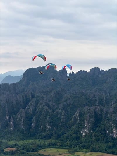 In Hong Ngern Village, Laos, three individuals are seen paragliding over a mountainous landscape under a cloudy sky. Each paraglider has a brightly colored wing, predominantly red, white, and blue. They are suspended from their respective paragliders by a harness and a motor. The mountains are covered in dense green foliage, with sharp, rocky peaks. In the foreground, a patchwork of agricultural fields is visible. The lighting suggests it is daytime, with diffused light from the overcast sky.