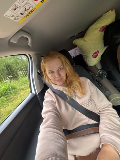 A young woman with blonde hair sits in the passenger seat of a car, looking at the camera. She is wearing a light pink hoodie and a seatbelt. Outside the car window, a lush green forest is visible, with a dirt road in the foreground. A stuffed toy and fishing rod are visible in the back seat. The car appears to be driving on a road in Kadochnik, Russia, as indicated by a previous description not present here.
