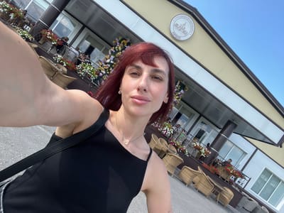 A woman with red hair takes a selfie in front of a building that appears to be a restaurant or cafe. The building has a light-colored facade with brown trim and windows. There are outdoor seating areas with wicker chairs and tables, some of which are occupied by people. Flower boxes filled with red and white flowers adorn the balcony and facade. A circular sign with text and an image of a building is mounted on the upper part of the facade. The sky is clear and blue. The location is identified as Akhlebaikha (Kovriginskii S/S), Russia.