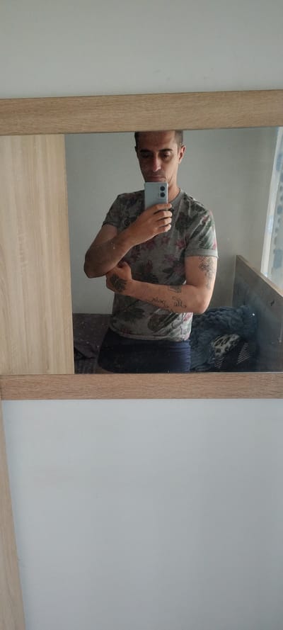 A person is taking a selfie in a mirror. The person is wearing a grey t-shirt with a floral pattern, and dark-colored pants. The person has tattoos on their arm. The mirror has a light wood-toned frame. The background shows part of a wall, a shelf, and some soft, possibly textured items on the shelf. No location information regarding Beguntsi, Bulgaria can be ascertained from this image.