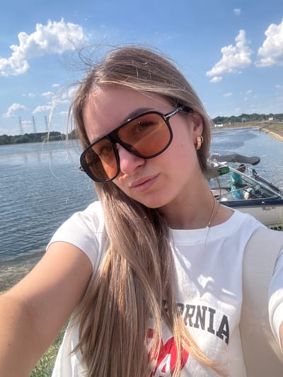 A young woman with blonde hair, wearing sunglasses and a white t-shirt, takes a selfie outdoors. She is positioned in the foreground, with a body of water, a boat with a person in it, and trees in the background. The sky is blue with white clouds. The woman is wearing a gold hoop earring. The t-shirt has "CALIFORNIA" and a red heart graphic visible. The scene appears to be a waterfront area.