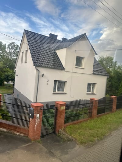 The image shows a two-story house with a dark grey tiled roof and white exterior walls. The house has several windows, some of which are covered with white curtains. A black fence made of vertical bars encloses the property, with red brick pillars at the gate and at intervals along the fence. The fence is fronted by a low red brick wall. To the left of the house, trees and greenery are visible. A small plaque with the number "3" is affixed to the front wall of the house. The ground in front of the house is paved with interlocking bricks. The sky above is partly cloudy with a mix of blue sky and white clouds. A power line runs diagonally across the upper part of the image. The scene appears to be located in a residential area, possibly in Połupin, Poland, given the context provided.