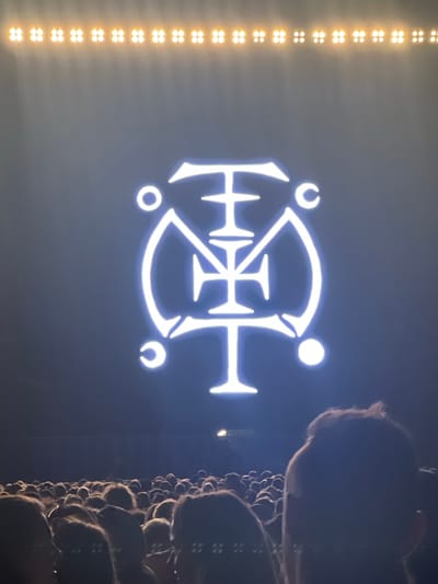 The image shows a concert audience in the foreground, with their backs to the camera, facing a large, illuminated symbol on a dark background. The symbol is white and appears to be a stylized geometric design with curves and sharp angles, resembling a crest or emblem. Above the symbol, a row of yellow lights is visible along the top edge of the screen. The audience is densely packed, with heads of various sizes and hairstyles filling the lower portion of the frame. The overall atmosphere suggests a live event or performance. The provided location, Pogórze, Poland, is not explicitly evident in the image.
