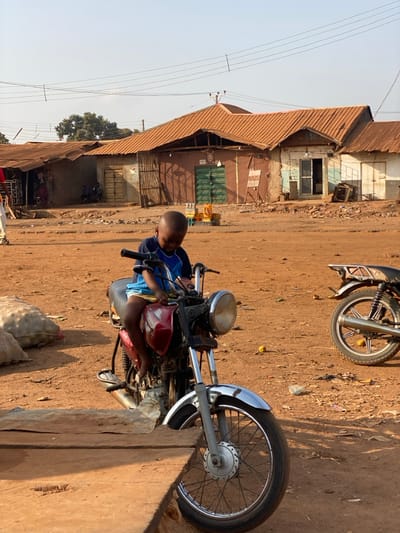A young boy sits on a parked red and black motorcycle in a dirt lot next to a row of aged buildings with rusty metal roofs. Another motorcycle is partially visible in the background. The buildings have patched walls and some closed shutters. Overhead power lines run along the street. The scene is bathed in sunlight, casting long shadows on the dusty ground.