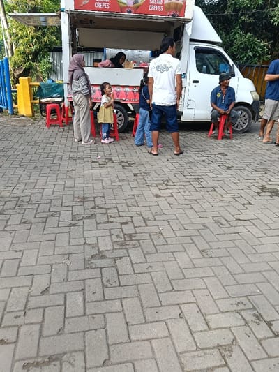 The scene depicts a gathering of people around a white food truck on a paved surface in Gembong, Indonesia. The ground is composed of gray interlocking pavers laid in a herringbone pattern, showing some wear and minor debris.
A white food truck, identified by prominent "CREPES" signage, is parked, with its serving window open. Behind the counter, a woman in a black hijab and pink top is visible, appearing to serve customers.
To the left of the serving window, a woman wearing a light brown hijab, striped gray and white long-sleeved shirt, and light brown trousers stands facing the truck. To her right, a young girl in a yellow dress and pink shoes stands on a red plastic stool, looking towards the truck and holding something to her mouth. Another child, dressed in a dark blue shirt and blue jeans, is partially visible behind the first child, also standing on a red plastic stool.
Centered in front of the truck's serving area, with his back to the viewer, stands a man in a white T-shirt with a dark graphic, dark blue shorts, and dark sandals.
To the right of the truck's front wheel, an older man sits on a red plastic stool, wearing a blue collared shirt, dark trousers, and a dark bucket hat, looking to his right. Partially visible on the far right, another man stands, wearing a dark blue shirt and light-colored shorts with sandals.
In the background, lush green foliage and trees are visible, along with a blue and yellow barrier or fence on the far left. Several red plastic stools are positioned near the food truck.