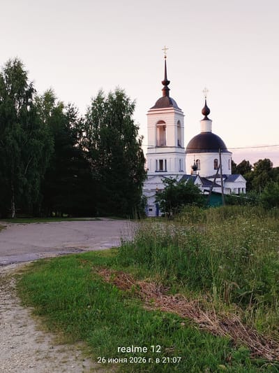 A white Orthodox church with a dark dome and a bell tower stands amidst lush green trees. The church has multiple arched windows and ornate crosses atop its spires. A dirt path leads towards the church from the lower left of the frame, bordered by tall grass and wildflowers. The sky is a pale pink and white, suggesting either dawn or dusk. The scene is peaceful and rural, likely in Novoe, Russia, given the timestamp "26 июня 2026 г. в 21:07" which translates to "June 26, 2026 at 21:07". There are no people visible in the image.