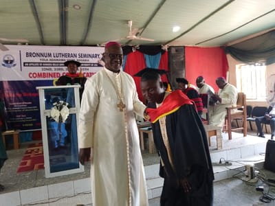The image shows a graduation ceremony taking place at the Bronnum Lutheran Seminary, Yola Adamawa State, Nigeria (located at 9.184, 12.522). The main subject is a man wearing a black graduation gown and a red sash, receiving a certificate from a man wearing a white robe and a gold cross necklace. Both men are standing on a stage, which is decorated with a large banner featuring the seminary's name and logo. The man in the white robe is reaching out to give the graduate a certificate, which is in a clear, framed case decorated with a white ribbon. There are other people sitting in the audience, including a man wearing a black gown and a woman wearing a red gown, and a man in a white robe. The room is decorated with red and blue curtains. The image is taken from a slightly low angle, looking up at the stage. The lighting is bright and the focus is on the two men on stage.