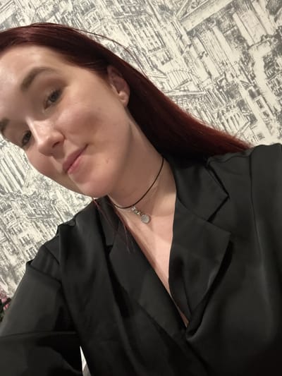 A young woman with reddish-brown hair and fair skin, wearing a black satin blazer and a choker necklace, is taking a selfie. She is positioned at an angle, with her face turned towards the camera. The background features a wall with a decorative pattern resembling a city skyline. The lighting is soft, highlighting her features and the texture of her clothing. No specific location within Pervouralsk, Russia can be determined from the image.