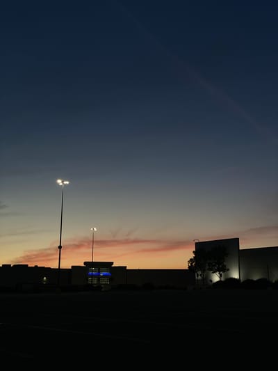 The image depicts a commercial building at dusk in Halfway, United States of America. The sky is a deep blue transitioning to lighter shades of orange and pink near the horizon. In the foreground, a dark, empty parking lot is visible. Two tall lampposts with multiple lights are illuminated. The building itself is silhouetted against the sky, with a section of its facade featuring bright blue illuminated signage that reads "VEETEN MALL". A single tree stands in front of another portion of the building, also silhouetted. The scene is devoid of people.