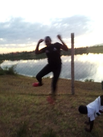 This image depicts an outdoor scene in Central Kasipul ward, Kenya, during what appears to be either dusk or dawn, indicated by the sky's light conditions. The foreground consists of dry, sparse grass and dirt. A chain-link fence, supported by at least one visible wooden post, traverses the mid-ground. Behind the fence, a body of water reflects the sky's light. Distant land or hills are faintly visible on the horizon beyond the water.
Two human figures are present. The primary figure, positioned towards the center-left, is blurred due to motion, suggesting an active pose such as jumping or dancing. This individual wears a dark-colored short-sleeved shirt and dark trousers. One foot, visible and raised, appears to be clad in red footwear. Both arms are bent at the elbows and raised, suggesting an upward or celebratory gesture.
A second figure is partially visible in the bottom right corner of the frame. This individual, possibly a child, is in a low, leaning posture, with their upper torso and head directed downwards and to the left. They are wearing a white short-sleeved shirt with faint markings. The figure's hands are obscured by the ground, indicating they might be resting on all fours or in a similar crouched position.