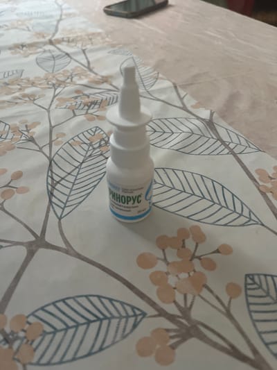 A white plastic nasal spray bottle, labeled in Cyrillic script, sits on a white tablecloth with a light brown and blue floral and leaf pattern. The bottle is upright, with its white cap on. The label on the bottle is partially obscured but the word "РИНОРУС" (Rinorus) is clearly visible. The tablecloth is laid out on a flat surface, possibly a table, and a corner of a mobile phone is visible in the upper right of the frame. The background is out of focus, but appears to be a room. The location is Түндүк айланып өтүүчү жол 84 га, Kyrgyzstan.