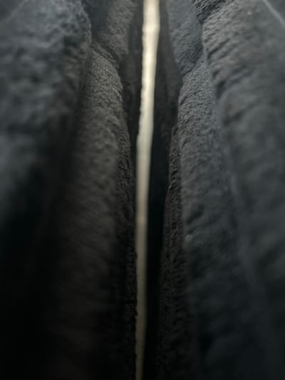 The image is a macro shot of a dark, fuzzy material, likely fabric, creating a narrow vertical gap with a bright light source visible in the center. The fabric appears to have a textured, slightly matted surface. The depth of field is shallow, with the edges of the fabric in sharp focus and the central gap and areas further away from the lens blurred. There is no discernible location or context within the city of Lynnwood, United States of America. The image focuses solely on the abstract texture and light interaction of the material.