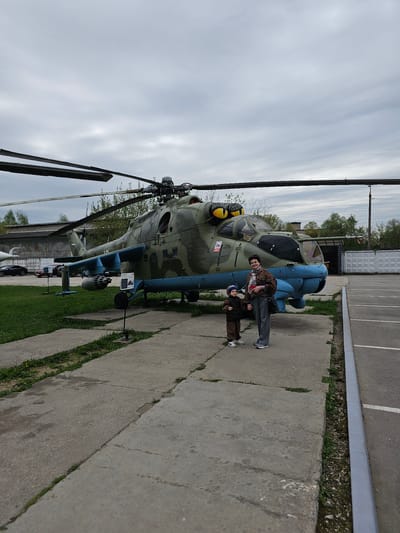 A man and a child are standing in front of a military helicopter. The helicopter is painted in shades of green and blue, and has its rotor blades extended. A building is in the background. The scene is located in Arkhangel’skoye, Russia.