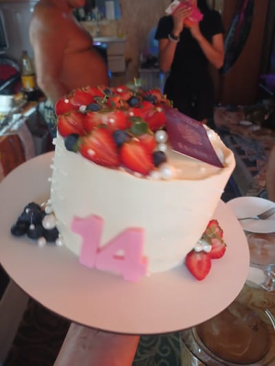 The image shows a cake decorated with strawberries, blueberries, and pearls. Two pink numbers, "14", are attached to the side of the cake. A passport is stuck into the top of the cake. In the background, a person in a black shirt is holding a pink object. The scene appears to be indoors, possibly a restaurant or a home. The overall impression is that of a celebration. The image is taken from a slightly high angle, looking down at the cake. The lighting is warm and appears to be artificial.