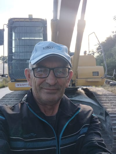 A man wearing a light blue baseball cap and eyeglasses is taking a selfie in front of a yellow excavator. He is wearing a dark jacket with blue and white stripes. The excavator has a yellow cab and large metal tracks. The background shows trees and a bright sky. The image appears to be taken in a rural or industrial area, possibly on the outskirts of Novoselytsia, Ukraine.