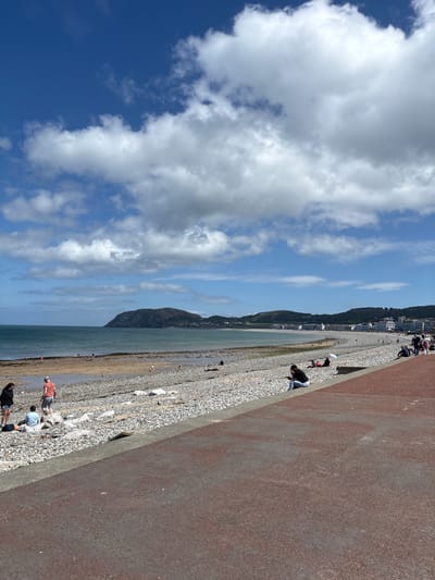 The image shows a pebble beach in Llandudno, United Kingdom, on a sunny day with a blue sky and scattered white clouds. The beach slopes down to the water on the left. People are scattered along the beach and the promenade, some bending down and others standing. A textured wall or barrier runs along the right side of the beach. In the distance, a mountain meets the water, with buildings nestled on the hillside.