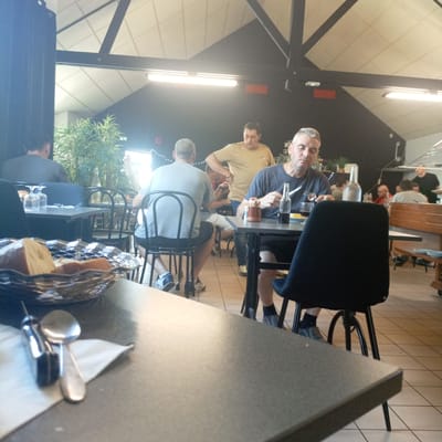 The image depicts an indoor dining setting, possibly a restaurant, with a high ceiling supported by dark beams. The lighting suggests it is daytime. In the foreground, a basket of bread and cutlery are visible on a dark table. Several people are seated at tables in the middle ground and background. One man in a blue t-shirt is seated at a table on the right, holding a fork and knife. Another man in a light brown t-shirt stands behind him. In the left middle ground, a person with their back to the camera is seated at a table. Further back, other patrons are visible, some with their backs to the camera. The environment appears to be a casual dining establishment. The scene is located in Derval, France.