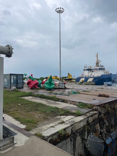 The image shows a port scene in Ebute Leki, Nigeria. A large blue and white ship, labeled "OCEANIC FRONTIER", is docked. Smoke is visible coming from the ship. Along the dock, several red and green buoys are stacked. A white truck and yellow cranes are visible in the background. A gray shipping container is in the foreground. A tall light pole with multiple lights is on the left. The sky is overcast. A concrete ledge runs along the edge of the dock, with some grass and vegetation growing near it. A metal grate covers a drain.