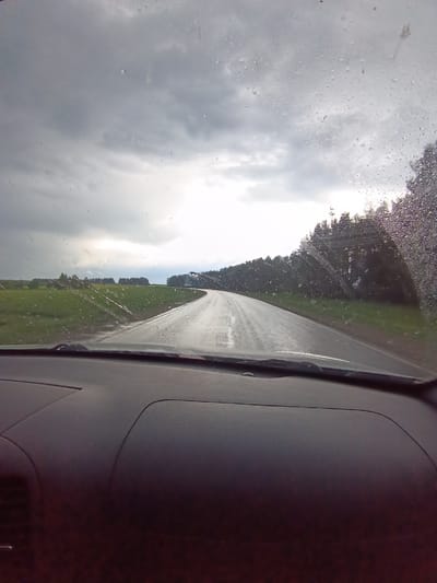The image is taken from inside a car, likely from the driver's perspective, looking out through the windshield on a rainy day. The road ahead is wet and winding, curving slightly to the right. Green fields are visible on the left side of the road, while a line of trees borders the right. The sky is overcast with gray clouds, and the windshield has water droplets obscuring the view. The dashboard of the car is in the foreground, dark in color and occupying a significant portion of the lower half of the image. There are no visible people or specific objects beyond the road, vegetation, and sky. The image provides no evidence to confirm a location in the city of Usola, Russia.