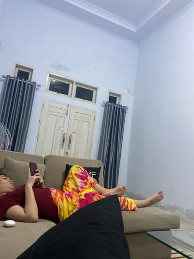 In a dimly lit room, a person reclines on a tan sofa, engrossed in their smartphone. They wear a maroon t-shirt and colorful tie-dye pants, with their feet bare and relaxed on the sofa. To their left, a white object, possibly an earbud case, rests on the cushion. Behind the person, a black pillow with a white design is visible, and a plush black cushion partially obstructs the foreground. The room features light blue walls with visible water damage and stains, and a white ceiling with decorative molding. Two sets of dark gray curtains frame windows, and a large white double door with red handles is situated in the background, with two smaller windows above it. A fan is partially visible on the far left. The scene appears to be within a residential setting in Walennae, Indonesia, given the typical architecture and interior design elements.
