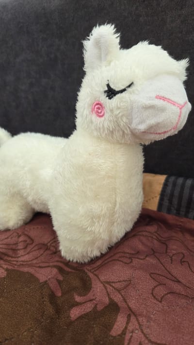 A close-up shot shows a white plush toy with pink and black embroidered details resting against a dark gray and patterned maroon background. The toy appears to be an animal with a closed eye, a pink cheek marking, and a small ear. The maroon background has a plant-like pattern in a darker shade of brown.
