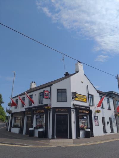 This is a full shot, eye-level, outdoor, daytime, vertical photograph of P.J. Bryson's Bar, a white building with black trim, located in Magherafelt, United Kingdom. The bar is situated on a corner, with two streets visible in the foreground. The building has a dark tiled roof and several windows. Red flags are flying from poles on the side of the building. A black sign with "P.J. BRYSON'S BAR" is visible above the entrance. A yellow sign with "OLD BUSHMILLS WHISKEY" is also displayed on the building. Two black barrels are placed outside the entrance. The sky is blue with some clouds, and a power line runs across the top of the frame. A street light and trees are visible on the left side of the image.