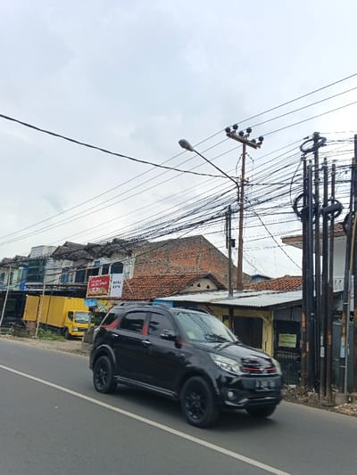 A black SUV moves along an asphalt road on an overcast day. Buildings and a yellow truck line the left side of the road. Overhead, a dense network of power lines crisscrosses above, supported by wooden and metal poles. Vegetation peeks out along the roadside, near a building with a corrugated metal roof. The overall scene depicts a typical urban or suburban street setting with a mix of residential and commercial elements.