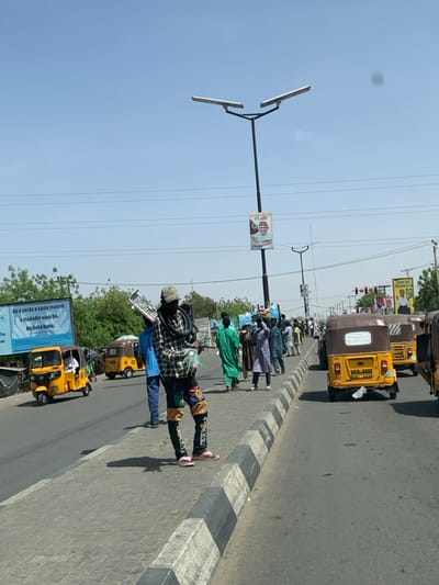 The image depicts a bustling street scene in Hotoro, Nigeria, under a clear blue sky. In the foreground, a man stands on a paved sidewalk, facing left. He wears a patterned bucket hat, a plaid long-sleeved shirt, dark pants with colorful patches and "SAX" lettering, and pink flip-flops. He carries a coiled black hose over his left shoulder, a white accordion-like object in his right hand, and a transparent plastic bag. The sidewalk features a black and white striped curb.
To the left, a multi-lane asphalt road runs parallel to the sidewalk. Two yellow Keke Napep tricycles are visible on the left side of the road, with one displaying "0171" and "KANO". A large blue billboard behind them features white text in Hausa: "Ba a yarda a kama mutum a madadin wani ba. Ba doka bane."
Further down the sidewalk, numerous pedestrians, many in traditional attire, walk in the same direction, some carrying items on their heads. On the right side of the road, a queue of yellow Keke Napep tricycles is present; the closest one shows a brown canopy and a license plate "KMC 3326".
Multiple black streetlights with solar panels are visible, one prominently in the midground, featuring a banner with a man's portrait and text including "ALH. MUHAMMAD ALLAH... A SADAUKI." Overhead power lines traverse the upper portion of the frame. Sparse trees and distant buildings complete the background. Strong shadows indicate bright daylight conditions.