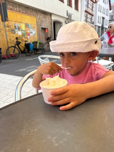 A child wearing a white hat and a pink shirt is seated at a dark-colored table, eating ice cream from a white paper cup. The child's right hand holds the cup while their left hand is near their mouth, possibly guiding the ice cream. An elderly woman with a red scarf and sunglasses is visible in the background near a white building with brown-trimmed windows. Behind the child and to the left, bicycles are parked against a yellowish wall covered in colorful posters. The location is likely in Riedlingen, Germany, given the architectural style of the building in the background.