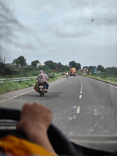 The image captures a view from inside a vehicle, looking out at a highway in Tandulwadi, Pr. Malkapur, India. A person's hand is visible on a steering wheel in the foreground. On the highway, a motorcycle with two riders, one operating the vehicle and another passenger, is traveling in the left lane. Both riders are covered with patterned cloths. Further ahead on the highway, several trucks are visible, including a large orange tanker truck in the left lane and a green truck with a blue tarp covering its load in the right lane. The roadside is lined with green vegetation and trees under a cloudy sky. Utility poles are also present along the side of the road.