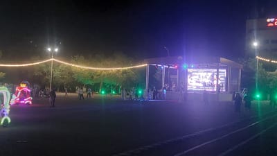 This nighttime outdoor image depicts a scene likely in Priozersk, Kazakhstan, featuring a temporary stage with a large LED screen broadcasting an event. The stage is illuminated with colorful lights, and several people are visible on and around it, some appearing to be performers or event staff. In the foreground, a group of people, including adults and children, are gathered, suggesting an audience or attendees. To the left of the stage, a brightly lit amusement ride or attraction, adorned with neon lights, is present, attracting attention. String lights draped between trees create a festive atmosphere. The environment appears to be a park or an open area within the city, with trees forming a backdrop and a building visible in the far right background. The ground is a dark, paved surface, possibly a recreational area or a street.