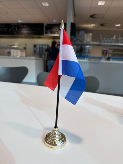 The image shows a close-up of a small Dutch flag on a table. The flag has red, white, and blue colors arranged in horizontal bands, with a white band in the middle. The flag is mounted on a thin black pole and stands in a gold, tiered base. The table is white and reflects the light. In the background, a blurred image of what appears to be a canteen or cafeteria can be seen. There are counters, chairs, and a person in the distance wearing a dark shirt and jeans. The overall impression is of a casual setting, possibly a break room or dining area. The specific location in Son en Breugel, Netherlands, is not discernible from the image itself, as there are no unique landmarks or identifying features of the town visible.