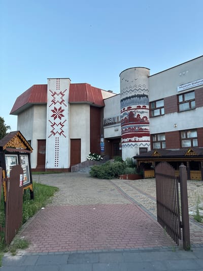 The image depicts the exterior of the Museum of Belarusian Culture, located in Olchowa Kładka, Poland. The museum building is a mix of architectural styles, featuring a white facade adorned with traditional red and white folk patterns, particularly a prominent star motif on one section and a cylindrical tower with a mural depicting rural scenes and nature. The building has a red, sloped roof. In front of the museum, there is a cobblestone pathway leading to the entrance, flanked by greenery and decorative wooden structures. A brown metal gate is visible in the foreground. The sky is clear and blue. No people are visible in the frame.