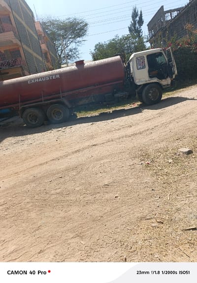 The image displays a static scene in Oloosirkon/Sholinke ward, Kenya, dominated by a white-cab, red-tank vacuum truck, commonly referred to as an "EXHAUSTER" vehicle, parked on an unpaved dirt road. The truck is positioned on the right side of the frame, facing towards the right, with the word "EXHAUSTER" clearly visible in white capital letters on its cylindrical red tank. The white cab exhibits signs of wear, and the passenger side door (right-hand drive orientation) appears to be missing a panel or partially open. The foreground consists of dry, light-brown dirt with distinct tire tracks and scattered small rocks and vegetation. In the background to the left, a multi-story building with light-colored walls and balconies featuring red railings is partially visible. To the right and behind the truck, an unfinished or dilapidated multi-story concrete structure with exposed rebar is obscured in part by dense green foliage and trees. Overhead, power lines are visible against a clear, light-blue sky. No individuals or active interactions are present in the scene.