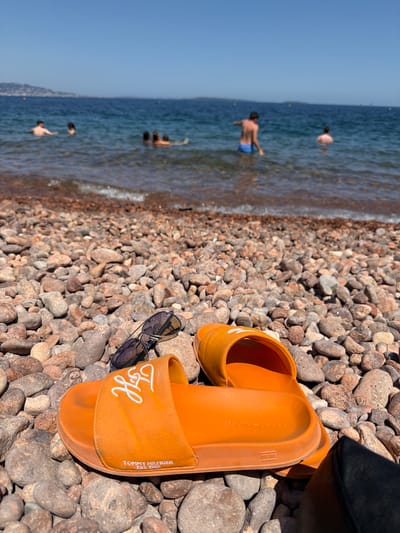 The image depicts a beach scene with a focus on a pair of orange Tommy Hilfiger slides and sunglasses resting on a pebble-strewn shore. In the background, several people are visible wading and swimming in the blue water of the Mediterranean Sea. A hazy coastline with distant hills is discernible on the horizon. The sky is clear and blue. This location is likely the beach of Théoule-sur-Mer, France, given its characteristic pebble beach and the view of the Esterel mountains across the bay.