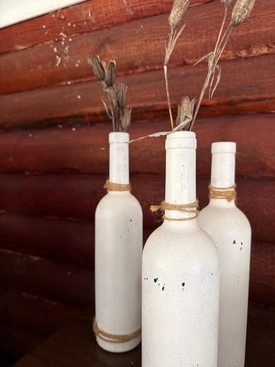 The image shows a still life arrangement of three white decorative bottles against a dark red wooden wall. The bottles are cylindrical with tapering necks and are adorned with rough twine wrapped around the neck of each. Each bottle contains dried, spiky plant stalks. The wooden wall has horizontal planks and a weathered appearance, with some white specks visible at the top. The lighting is diffused, casting soft shadows. The scene appears to be indoors or in a covered outdoor area. The location is specified as Nagornyi, Russia.