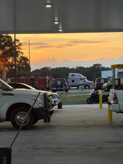 The image depicts a gas station scene at sunset in Piedmont, United States of America. A white truck is parked in the foreground, partially obscured by a canopy with overhead lights. To the right, another white truck is visible, as well as a black motorcycle. In the background, several other vehicles are parked, including a purple truck, a red truck with a load, and a large grey semi-truck. A stop sign is prominently displayed on a pole behind the red truck. The sky is illuminated by the warm orange and yellow hues of the setting sun, with fluffy clouds scattered across it. Trees line the horizon behind the gas station. The ground is paved, with yellow poles marking the parking or fueling spots.