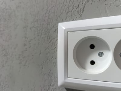 A close-up, eye-level, medium shot shows a white electrical outlet mounted on a textured gray wall. The outlet has two circular openings and a central screw. The wall's surface is rough and uneven, with visible indentations and raised areas. The lighting appears to be artificial, casting subtle shadows on the wall's textured surface. The image is cropped to focus on the outlet and a portion of the wall. The scene is indoors, possibly in a residential or commercial building. The location in Uchquduq, Uzbekistan is not discernible from the visual information provided in the image.