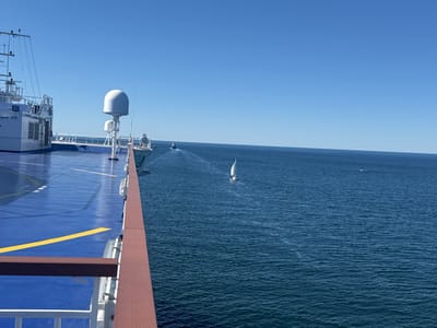 The original image shows a view from a large white ferry, likely the Stena Danica, with its blue deck and yellow lane markings visible in the foreground. The ferry is at sea under a clear blue sky. In the distance, a small white sailboat is visible, navigating the deep blue ocean. Further in the distance, a smaller boat leaves a wake behind it, also heading towards the horizon. The location is implied to be near Drottningskär, Sweden, suggesting the ferry is traversing the waters of the Baltic Sea. There are no people visible in the image. The overall scene is one of a calm, sunny day at sea.