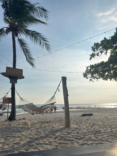 The image shows a hammock strung between two wooden posts on a sandy beach in Santa Teresa, Costa Rica. The hammock is white and has a woven pattern. There are several palm trees in the background. The sky is blue and the sun is shining. There is a sign that reads "MANZU" on a post. It appears to be a restaurant or bar with string lights strung overhead, and there are people in the distance on the beach. The location of this scene is on the beach at latitude 9.635, longitude -85.161.
