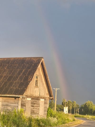 In Saulgoži, Latvia, a weathered wooden barn stands under a dramatic sky. The barn has a corrugated metal roof and wooden plank siding, with two boarded-up window openings on the lower section and a single small window on the upper gable. Tall green grass and some wildflowers grow in front of the barn. To the right, a faint rainbow arcs across the dark, cloudy sky. A simple wooden utility pole with wires is visible near the barn, and beyond that, a line of trees and more utility poles form the horizon. A paved road curves into the distance on the right side of the frame.