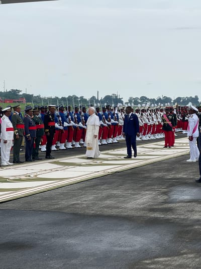 This image captures a formal outdoor military ceremony in Malabo, Equatorial Guinea, likely a state reception or review of troops. In the foreground, a prominent figure in white religious vestments, consistent with Pope Francis, stands on a patterned carpet, facing a long line of military personnel. Adjacent to him, slightly behind, is a man in a dark blue suit.

The military personnel are meticulously arranged in multiple rows, extending into the background, all standing at attention. They are dressed in diverse ceremonial uniforms, including distinctions of red trousers with blue jackets, dark green/black uniforms, and white uniforms with sashes, many carrying rifles.

The ground consists of a dark asphalt or concrete surface, partially covered by an ornate, light-colored patterned carpet where the main figures are positioned. The background features a horizon line dominated by lush green trees and glimpses of distant structures or buildings. The sky is uniformly overcast, indicating daytime with diffused natural light and no harsh shadows. The overall scene conveys a sense of solemnity and discipline befitting an official ceremonial event. No legible text is visible in the image.