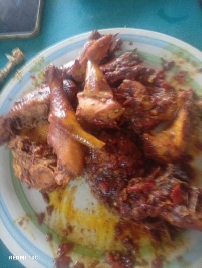 A plate of cooked chicken pieces heavily coated in a reddish sauce, possibly chili-based. The chicken varies in size, with some drumsticks and other portions visible. The plate has a white base with concentric blue and green rings around the edge. Some yellow liquid or sauce is visible on the plate. A partial image of a cell phone appears in the top left corner. Bone of the chicken can be seen.