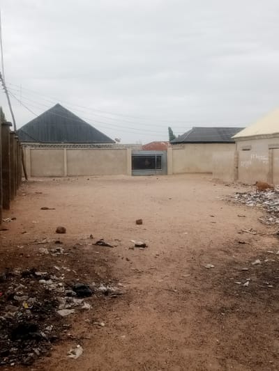 The image depicts a trash-strewn, undeveloped plot of land in what is purportedly the city of 0801712591, Nigeria. The ground is reddish-brown soil, sparsely covered with debris. Litter, including plastic, paper, and general waste, is scattered across the foreground and appears concentrated in a large pile at the bottom left corner.
A gated concrete wall forms the rear boundary of the property. Barbed wire is visible atop the wall. Adjacent buildings are visible beyond the wall.
To the right, a building with graffiti-covered exterior is present. A goat is visible near the building. The sky is overcast, suggesting poor weather conditions. Power lines are visible overhead.