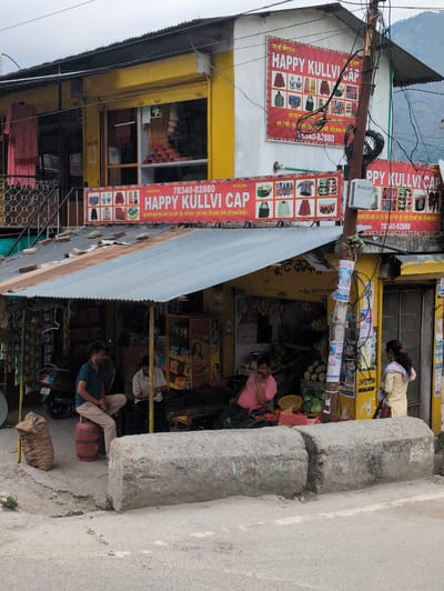The image shows a street scene in Bajaura, India, featuring a small shop with a yellow facade and a corrugated tin roof. A large red sign with the text "HAPPY KULLVI CAP" and a phone number is prominently displayed on the wall of the building. Below this, another banner with similar text and images of clothing items is visible.
In front of the shop, under a covered area, several people are present. One man is sitting on a red gas cylinder, wearing a blue shirt and light-colored pants. Another man is seated behind a counter, dressed in a light-colored shirt. A woman in a light-colored outfit is standing near the entrance of the shop, looking inside.
The environment includes a variety of goods displayed for sale, such as produce like bananas and vegetables, as well as packaged items. A scooter is parked near the shop entrance, and a large sack is placed on the ground. The street is paved, and concrete barriers are visible in the foreground. The overall scene depicts a typical commercial setting in a small town.