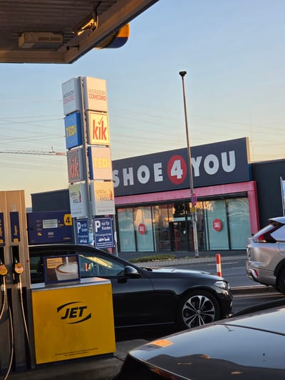 The image depicts a street scene in Bischofsheim, Germany, likely at a gas station and retail area. In the foreground, a yellow JET gas pump is visible, with fuel hoses attached. Next to it, a black car is parked. Behind the car and pump, a row of storefronts with prominent signage is present. One building displays the large white text "SHOE 4 YOU" with a red circle containing a "4" in the center. Red "SALE" signs are affixed to the windows of this establishment. To the left of "SHOE 4 YOU," a tall sign tower displays logos for various businesses, including "MATRATZEN CONCORD," "TEDI," and "kik." Overhead, power lines crisscross the sky. A street lamp stands tall to the right of the sign tower. A silver SUV is partially visible to the right of the "SHOE 4 YOU" building. The overall scene is captured under a bright, possibly late afternoon or early morning sky.