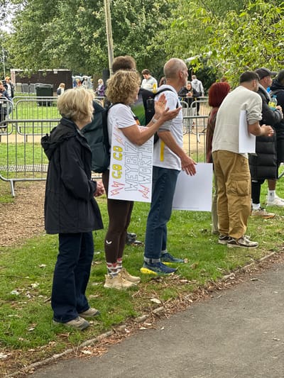 This image captures an outdoor public gathering in Weymouth, United Kingdom. The foreground presents a line of individuals standing on a grass verge, oriented towards the right side of the frame, with a paved edge delineating the bottom of the scene. From left to right: an older female with short blonde hair wears a dark, possibly black, long-sleeved jacket and dark trousers. Next, a female with curly light brown hair wears a white t-shirt, dark brown leggings, and a dark backpack. Affixed to her back is a white sign with hand-drawn red and yellow text: "GO GO GO WEYBIZ WORTH," accompanied by lightning bolt graphics. Her hands are raised and clasped in a clapping motion. Adjacent to her, a bald male in a light grey t-shirt, blue jeans, and blue athletic shoes also has his hands raised, and a plain white rectangular sign is tucked under his left arm. To his right, a female with reddish-brown hair is partially visible from behind, followed by a male in tan cargo pants and a light-colored long-sleeved shirt, holding another plain white sign against his left side. At the far right, a male wearing a dark puffer jacket and holding a clear plastic bottle is partially visible.
In the mid-ground, a series of interconnected metal crowd control barriers stretches horizontally across the frame, separating the foreground subjects from the background. The background features a dark grey cylindrical bin, a small brown utility building, and a dense canopy of green foliage from mature trees, indicating a park or open green space. The scene is illuminated by natural daylight, suggesting an overcast condition due to the soft light and lack of harsh shadows.