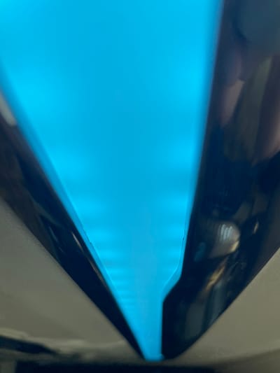 The image shows a close-up, abstract view of a bright cyan light emanating from what appears to be the interior of a device. The light is diffused and forms a V-shape, with dark, reflective surfaces on either side. The surfaces have a glossy finish, and subtle reflections of what could be a human hand and some internal components are visible. The overall impression is of a high-tech or industrial setting, but the specific location within Dong Nai City, Vietnam, cannot be determined from the visual information provided.